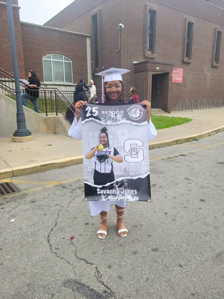 Savanna Jones in a white graduation gown and cap, holding a senior banner with her image in a softball uniform.