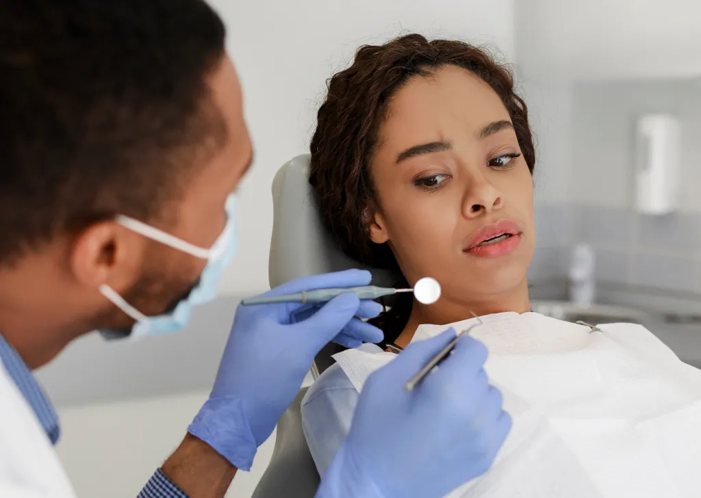 A scared Black woman looks fearfully at dental tools held by a masked dentist in a clinic.