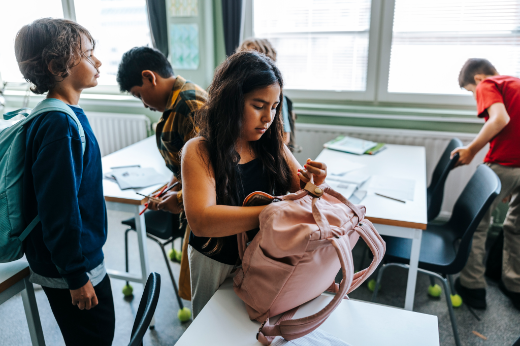 A schoolgirl packs her bag while standing in a classroom.