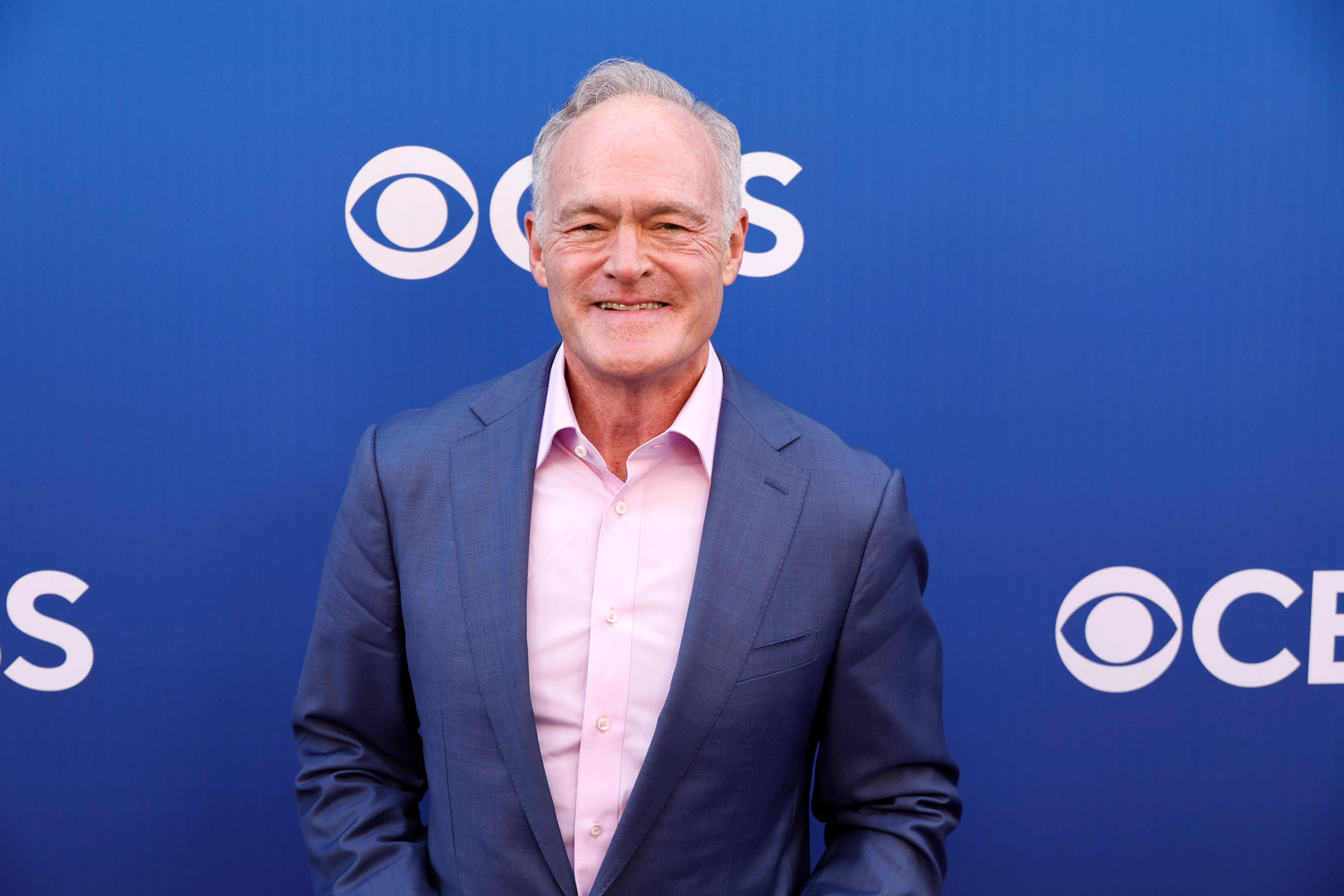 Scott Pelley wearing a blue suit and pink shirt in front of a CBS backdrop.