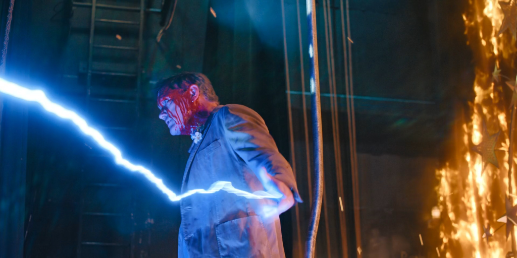 Matthew Lillard with a bloodied face and suit, holding a bolt of blue lightning, with flames in the background.