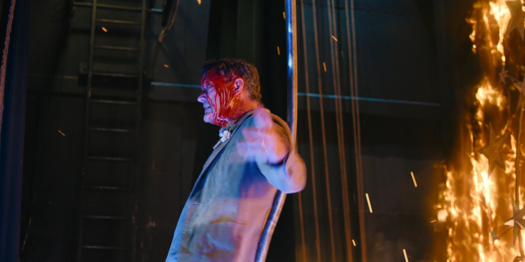 Matthew Lillard with blood on his face standing in front of a fire.