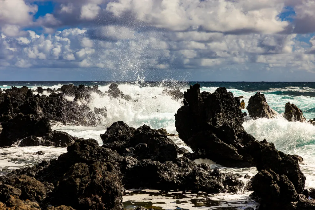 Waves crashing against the rugged black lava rock coastline of Maui, Hawaii, under a dramatic cloudy sky.