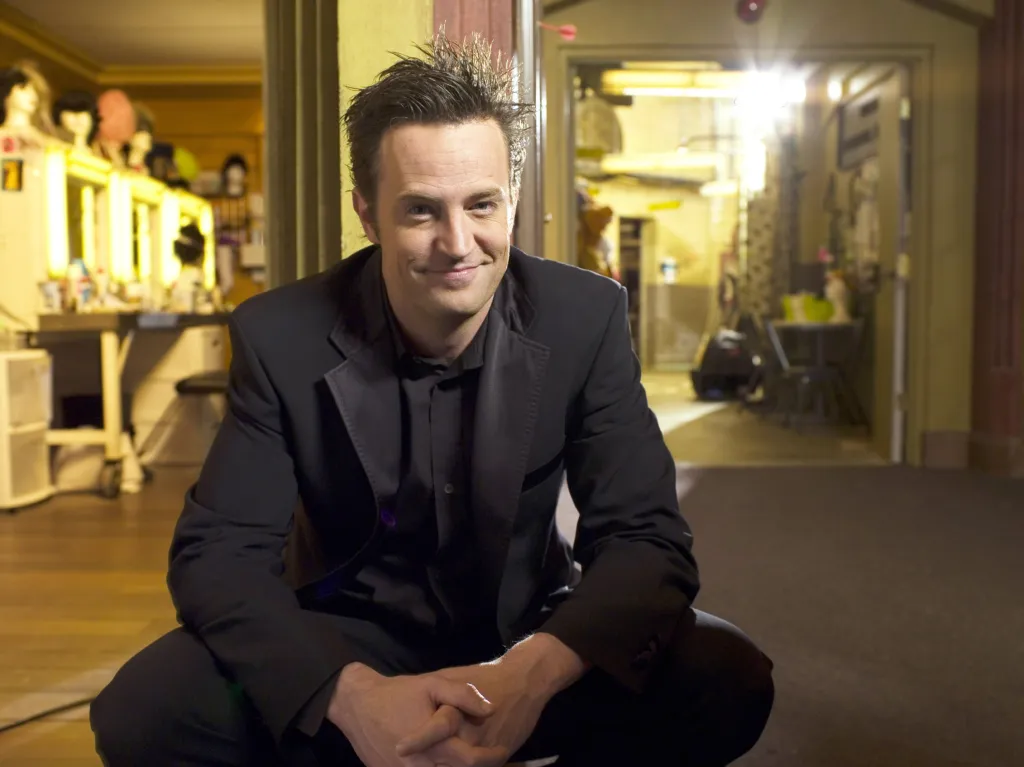 Actor Matthew Perry squatting in a hallway wearing a black suit and shirt in a promo photo for Studio 60 on the Sunset Strip.