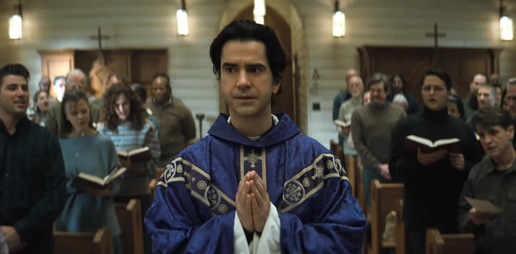 Hamish Linklater as Father Paul in Midnight Mass, standing in a church with his hands clasped in prayer before a congregation holding hymnals.