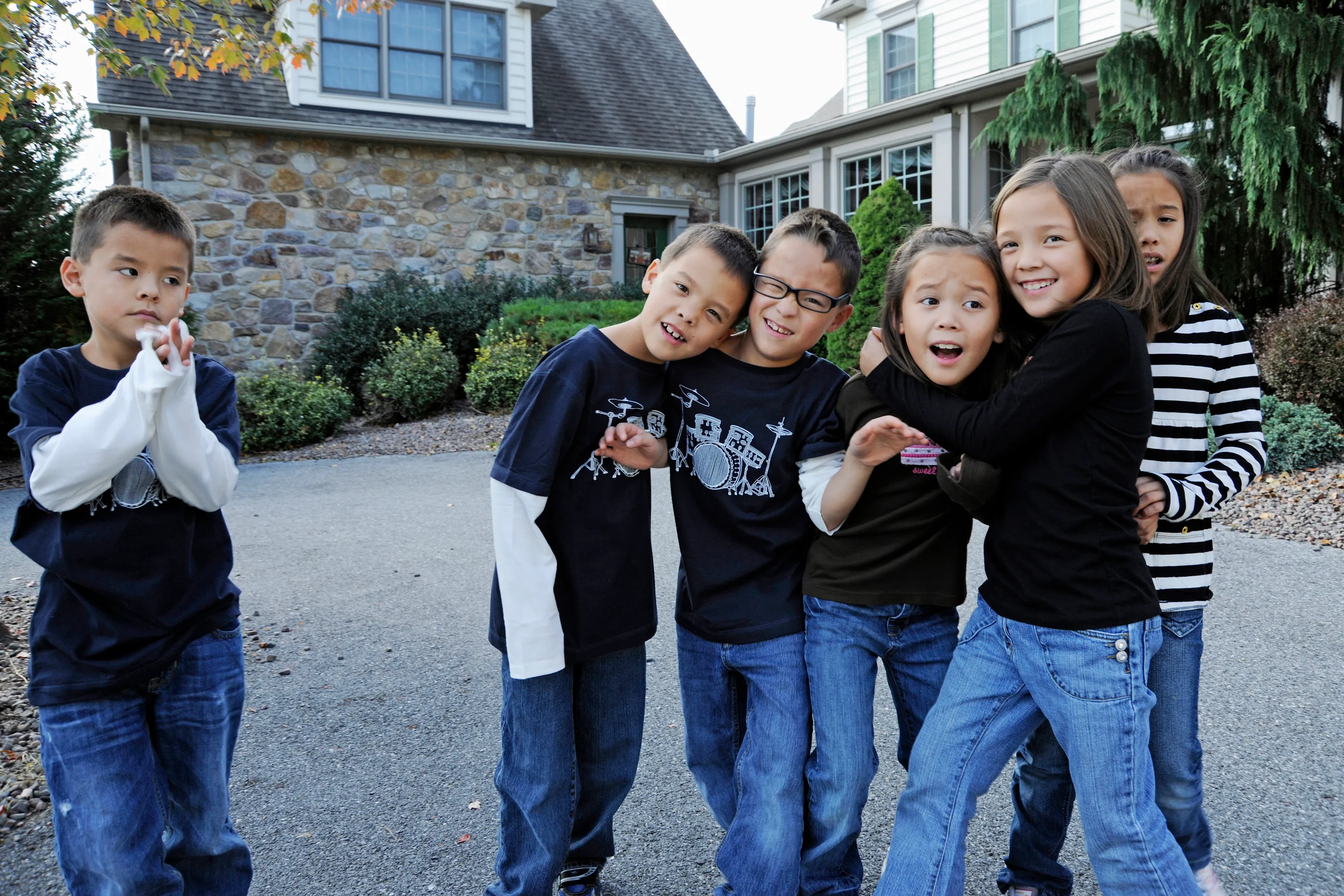 Six children pose outside a house, with one boy standing separately to the side, looking skeptical.