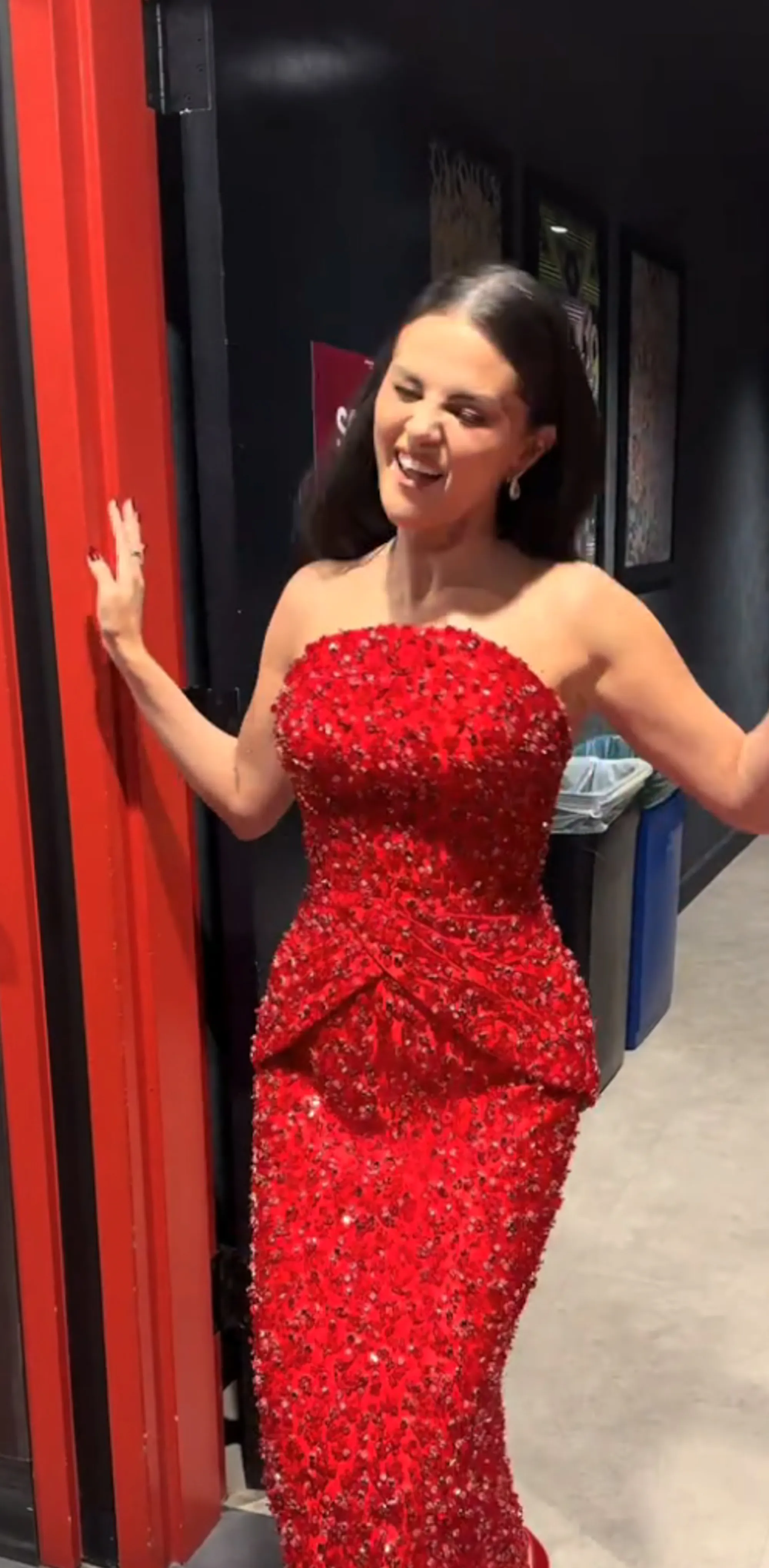 Selena Gomez wearing a red strapless dress, laughing while standing in a doorway.
