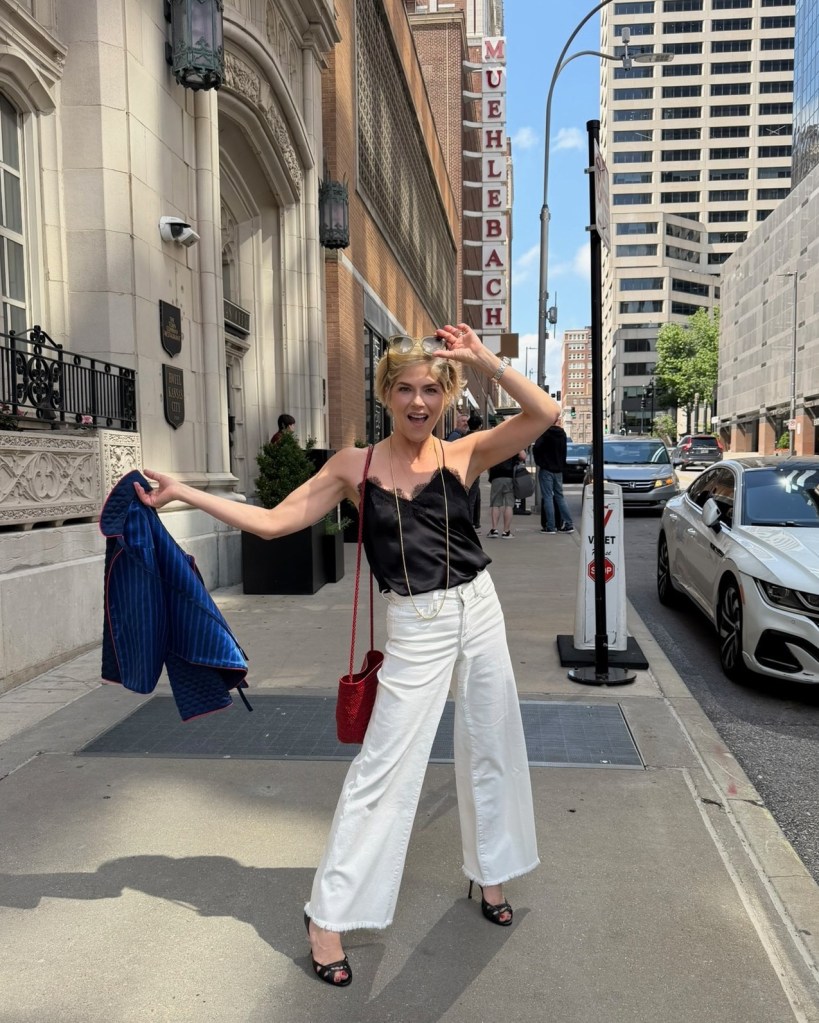 Selma Blair stands on a city sidewalk, wearing a black camisole, white wide-legged pants, and sunglasses, holding a blue jacket in one hand and smiling.