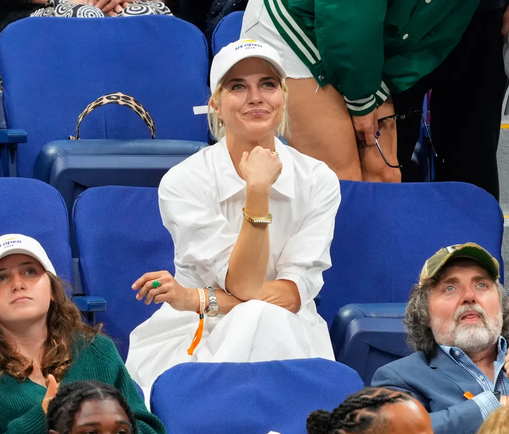Selma Blair sitting in the stands at the US Open, wearing a white dress and baseball cap.
