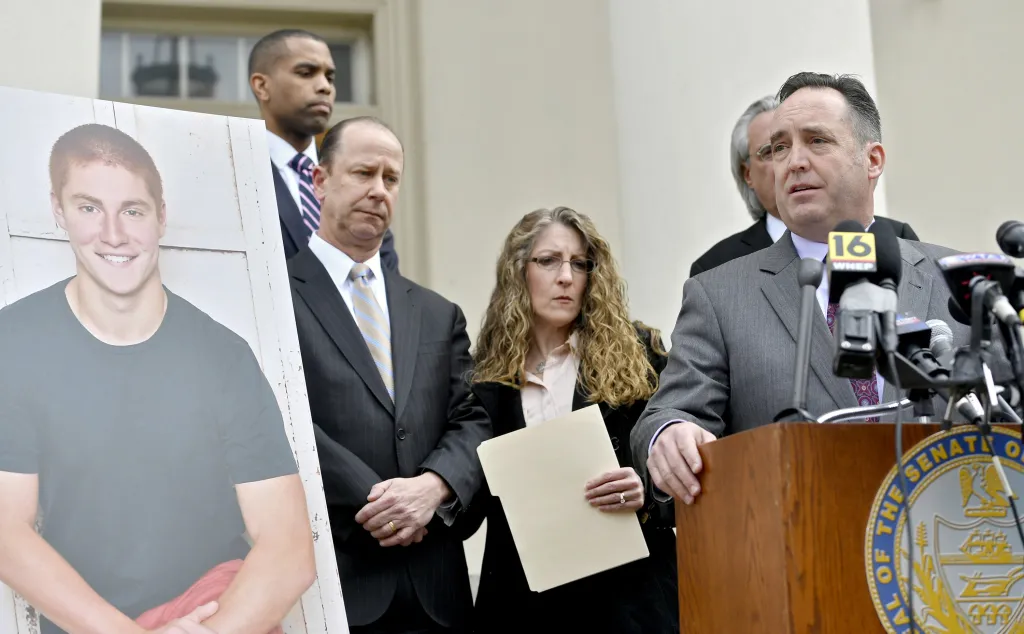 Senate Majority Leader Jake Corman speaks at a podium to introduce anti-hazing legislation named after Timothy Piazza, whose portrait stands to the left, alongside Jim and Evelyn Piazza.