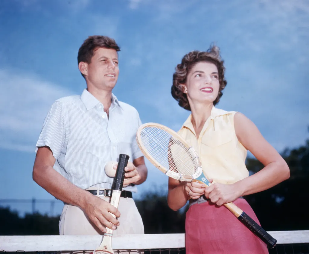 Senator John F. Kennedy and his fiancée, Jacqueline Bouvier, holding tennis rackets on a tennis court.