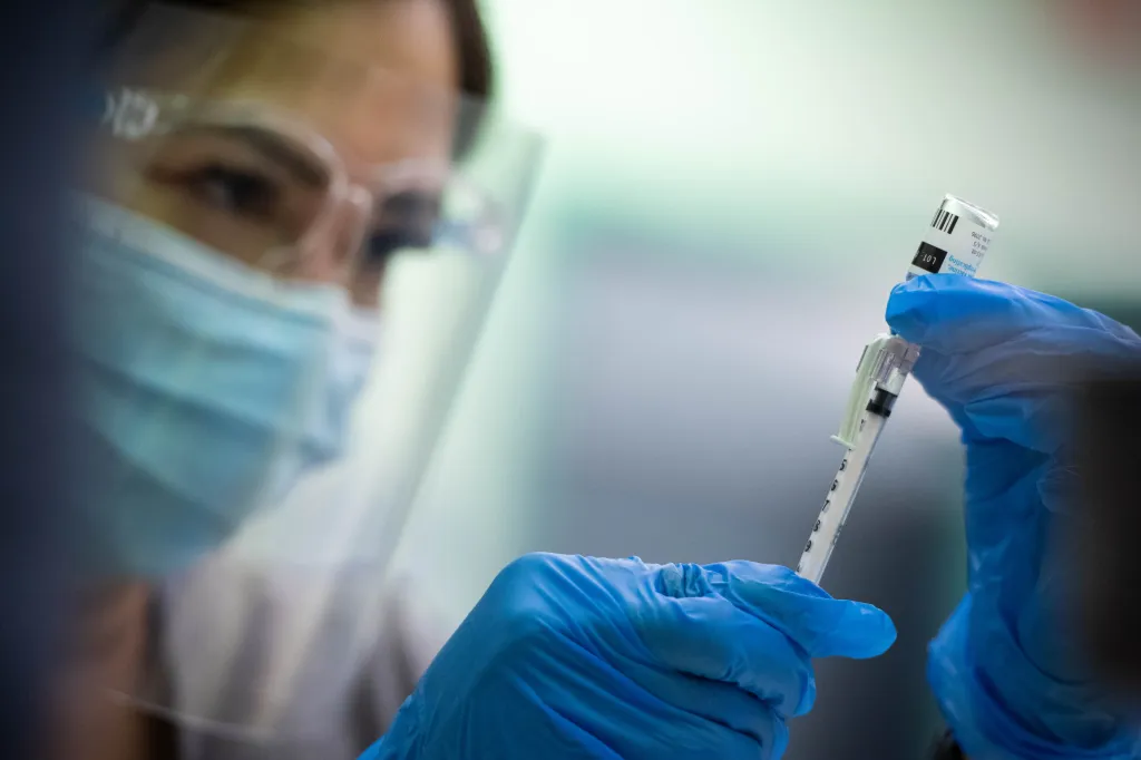 An LVN wearing a face mask and face shield draws a vaccine dose from a vial with a syringe.