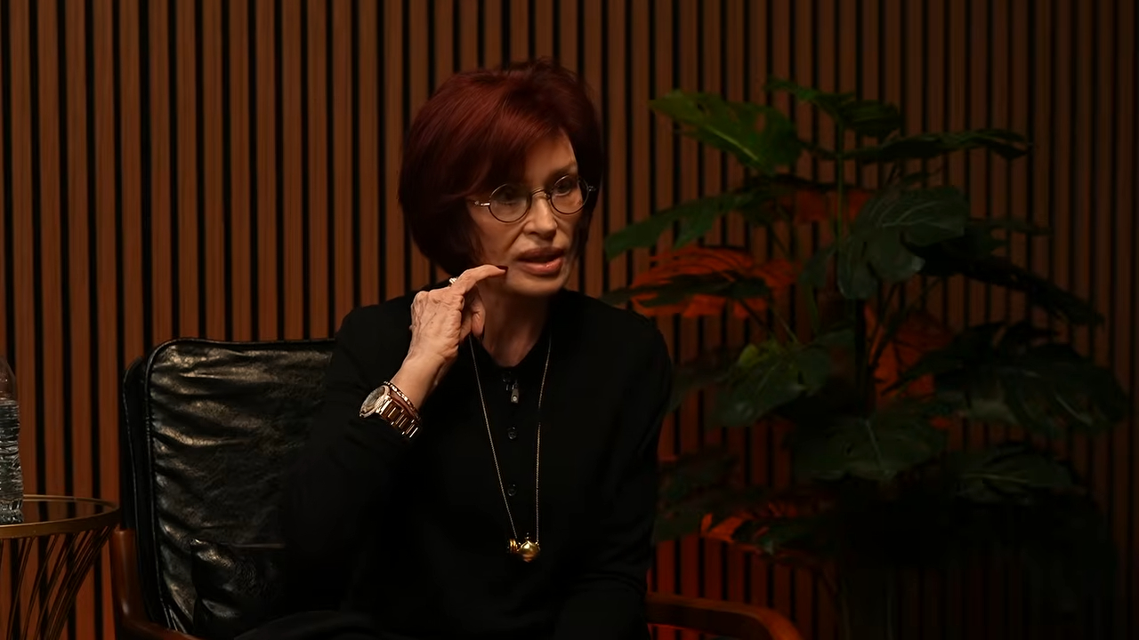Sharon Osbourne sitting in a chair and talking, with one hand touching her cheek.