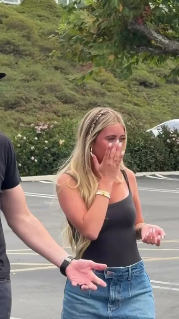 A teenage girl reacts with excitement and covers her face while standing in a parking lot next to a man.