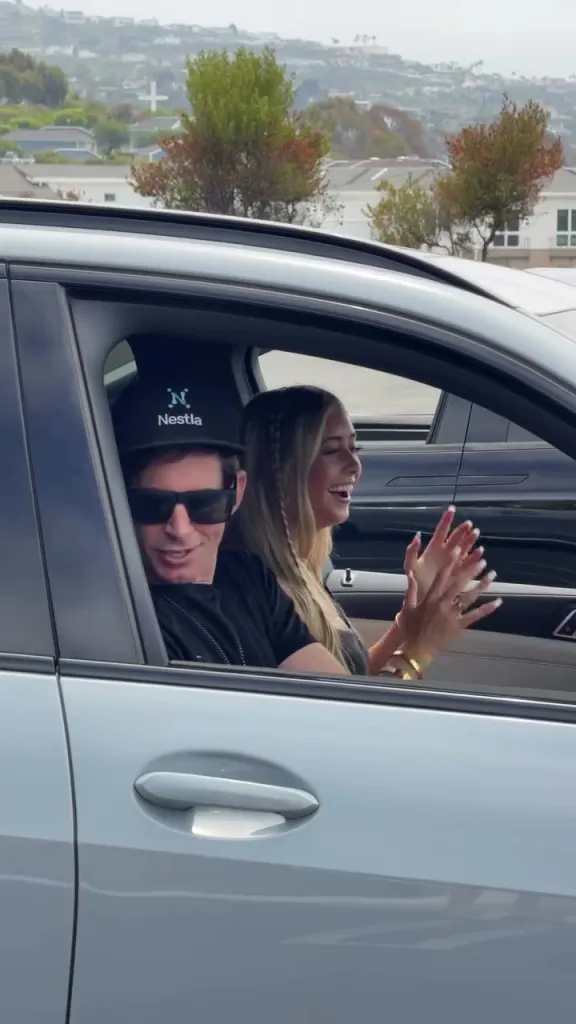 A man in sunglasses and a young woman sitting in a car, smiling and clapping.