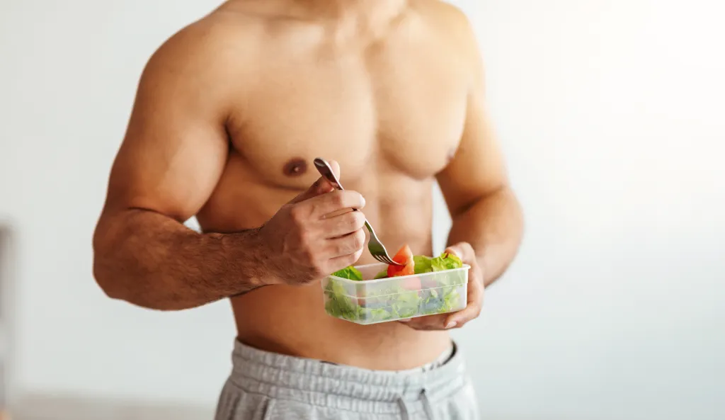 A shirtless man eats a healthy vegetable salad from a container in a bright kitchen.