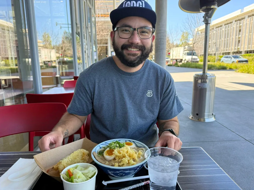 Jonathan McKinsey sitting at an outdoor restaurant table, smiling at the camera with a bowl of ramen and side dishes.