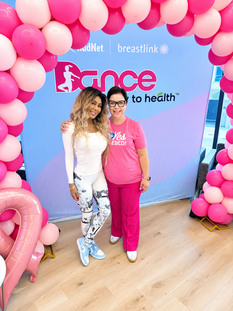 Two women standing in front of a blue backdrop with pink balloons at a