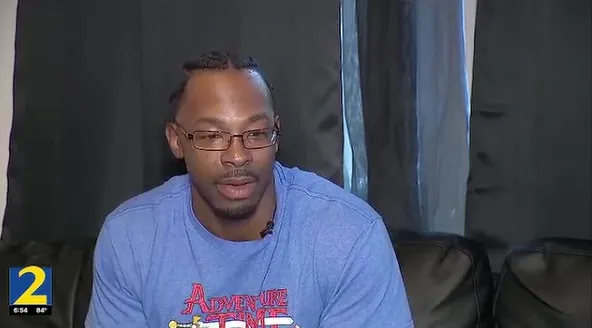 A Black man wearing glasses and a blue shirt sitting in front of dark curtains.