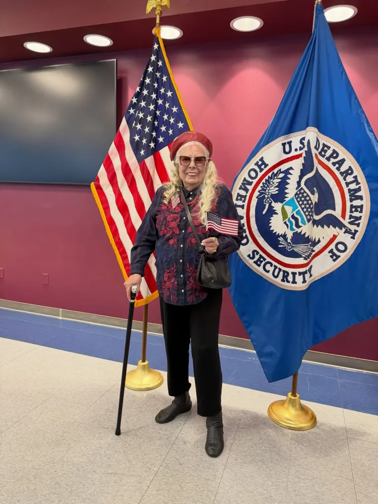 Joni Mitchell holding an American flag between U.S. and Department of Homeland Security flags after becoming a U.S. citizen.
