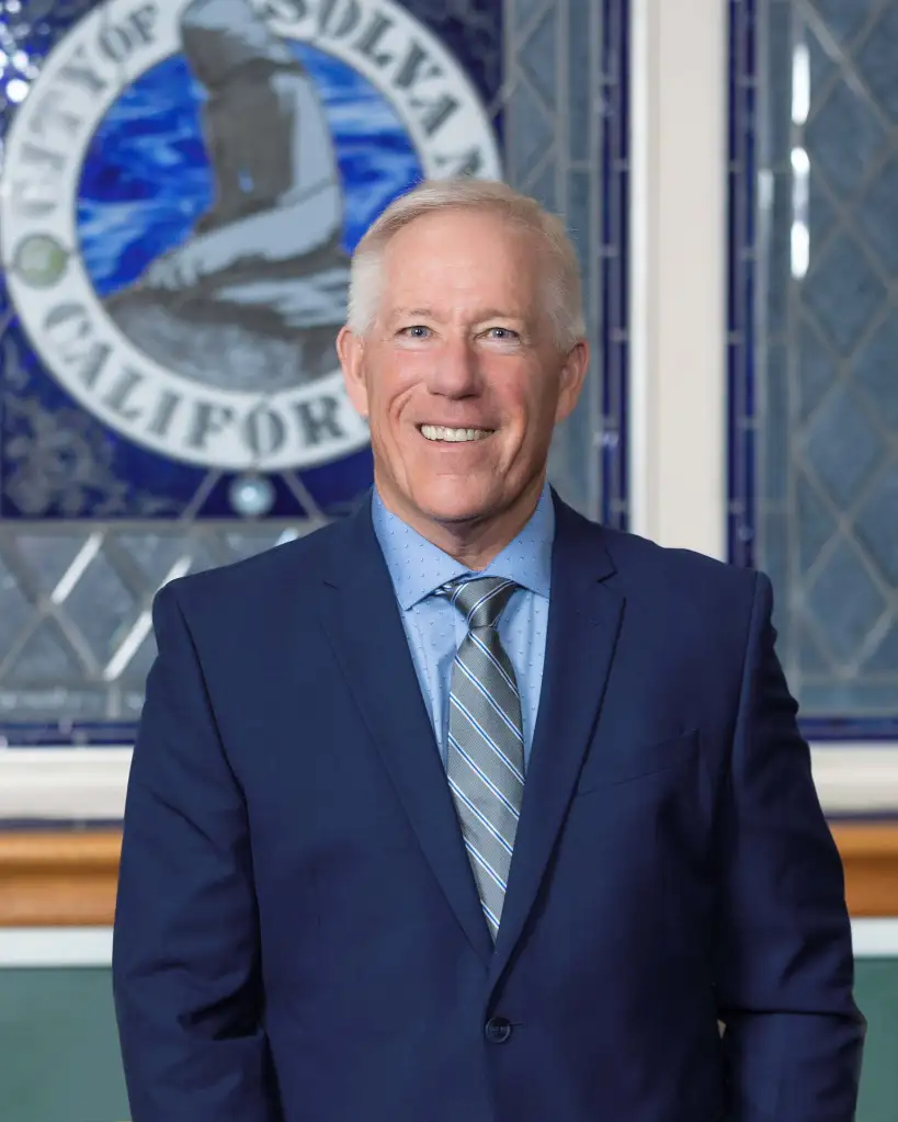 Solvang Mayor David Brown wearing a blue suit and striped tie in front of the city seal.