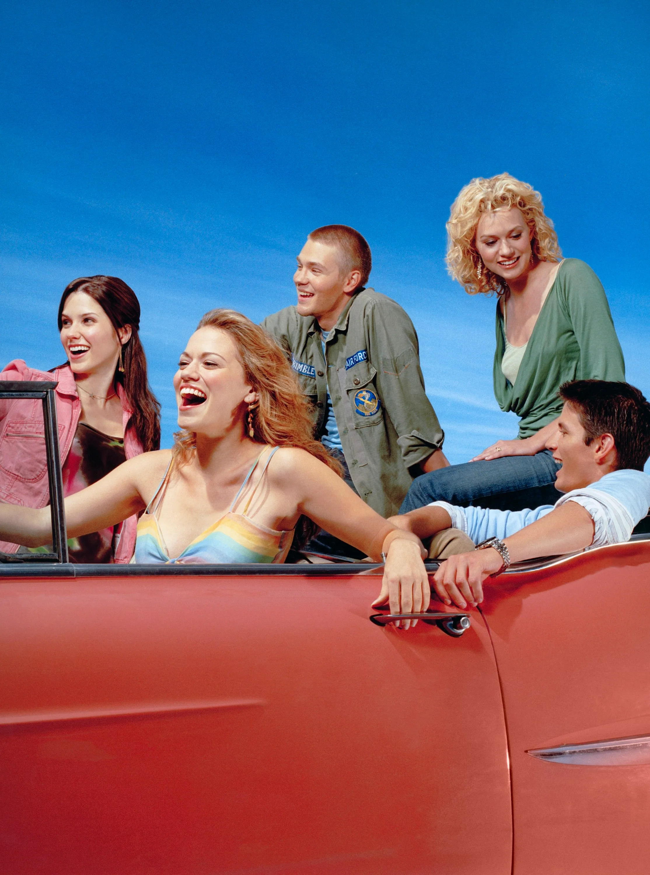 Cast of One Tree Hill laughing in a red convertible against a blue sky.