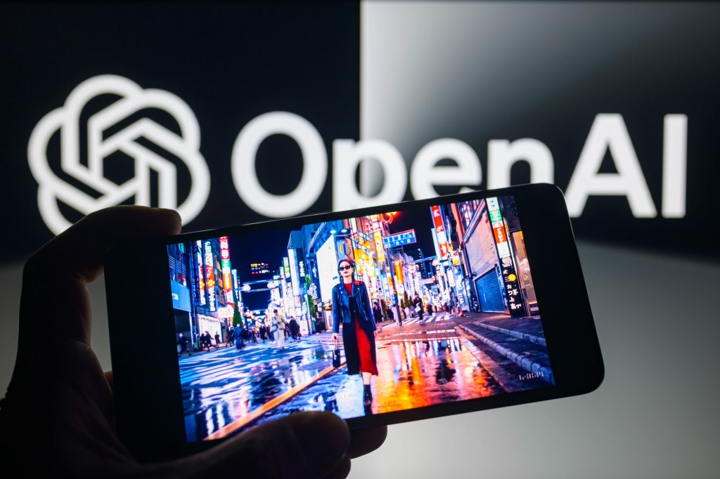 A hand holding a smartphone displaying the Sora text-to-video model, with the OpenAI logo in the background.