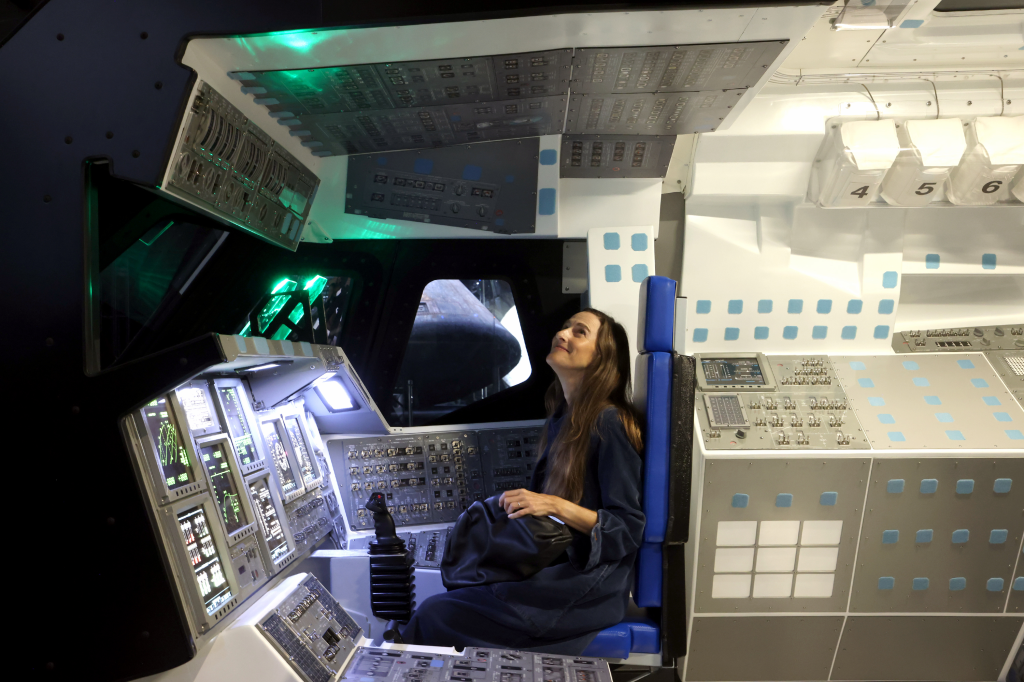 A woman sits in the cockpit of the Space Shuttle Endeavour display at the Samuel Oschin Air and Space Center, looking up with a smile.