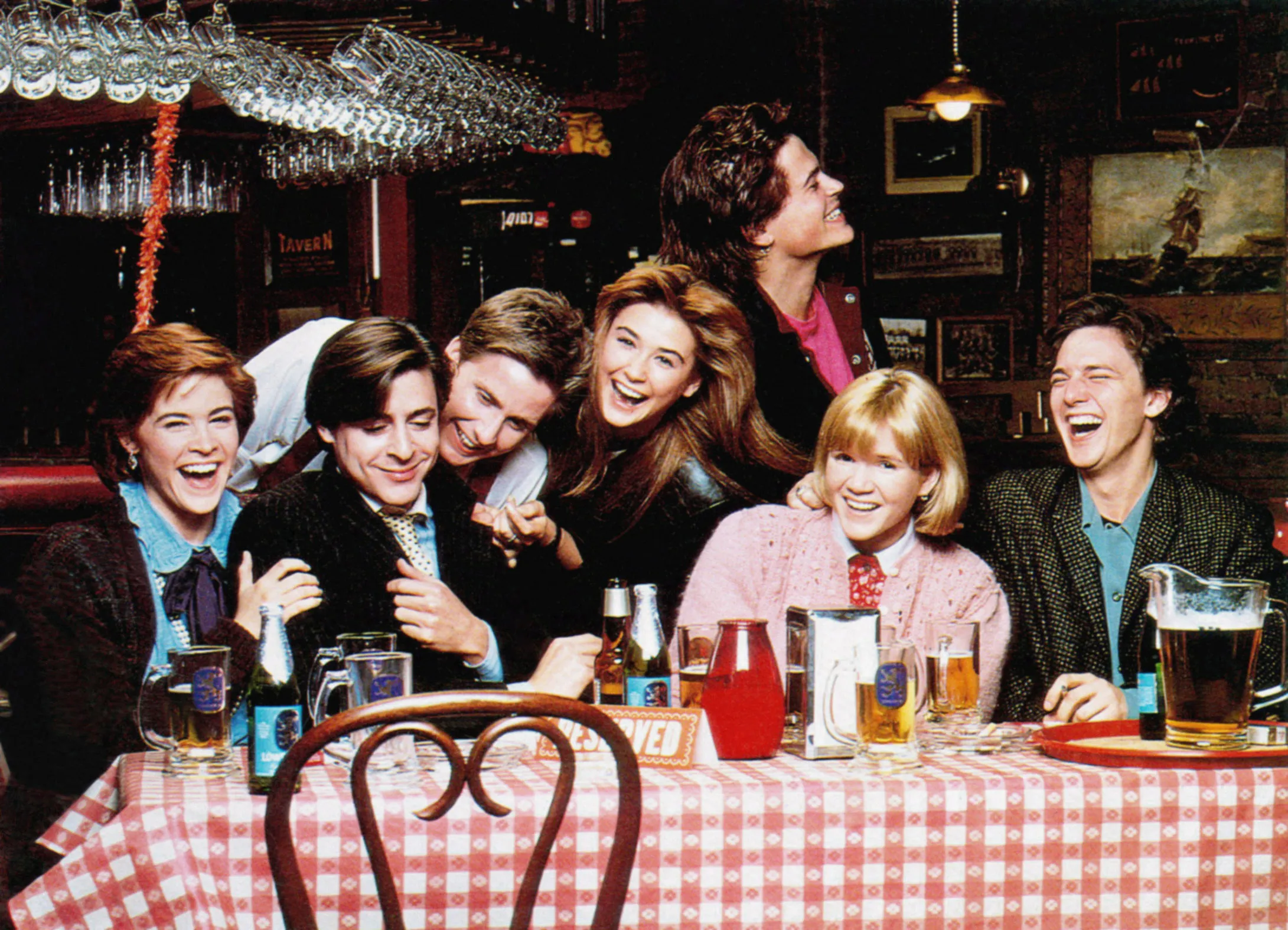 Cast members of St. Elmo's Fire, including Ally Sheedy, Judd Nelson, Emilio Estevez, Demi Moore, Mare Winningham, Rob Lowe, and Andrew McCarthy, gathered around a table in a pub.