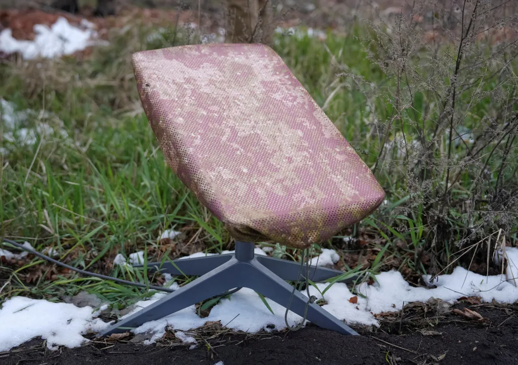 A Starlink satellite internet system with a camouflage cover set up on a stand in a snowy, grassy area.