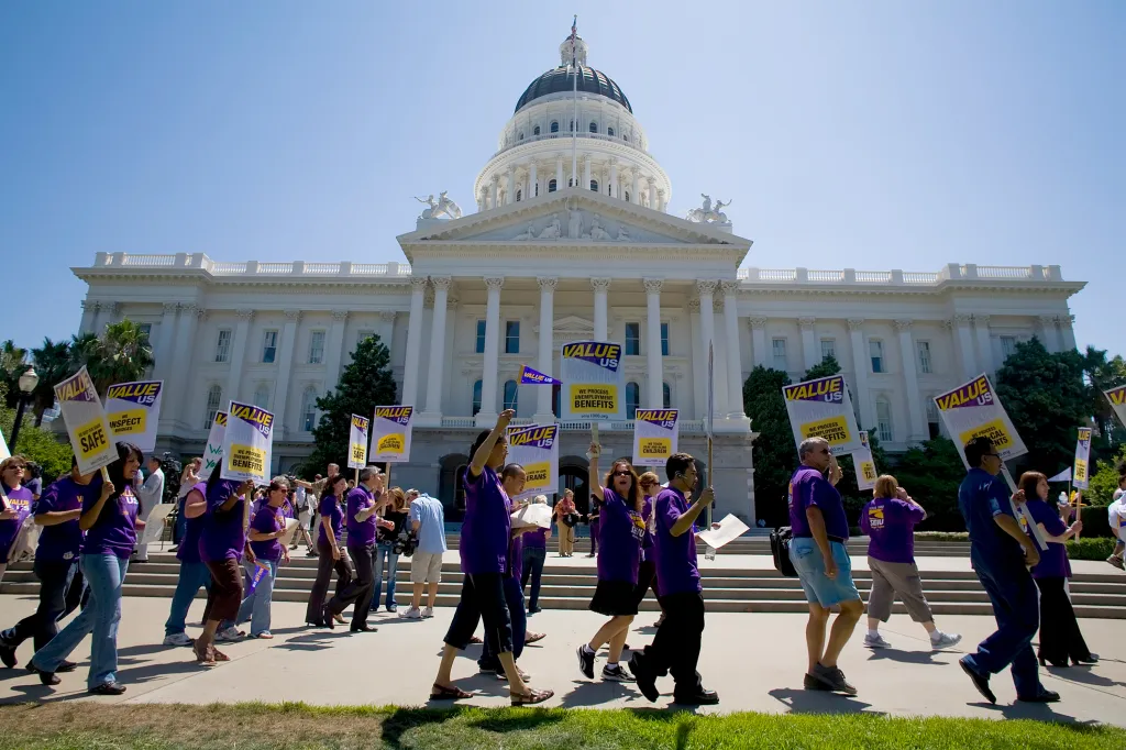 State workers protest in front of the California State Capitol, holding signs that read 