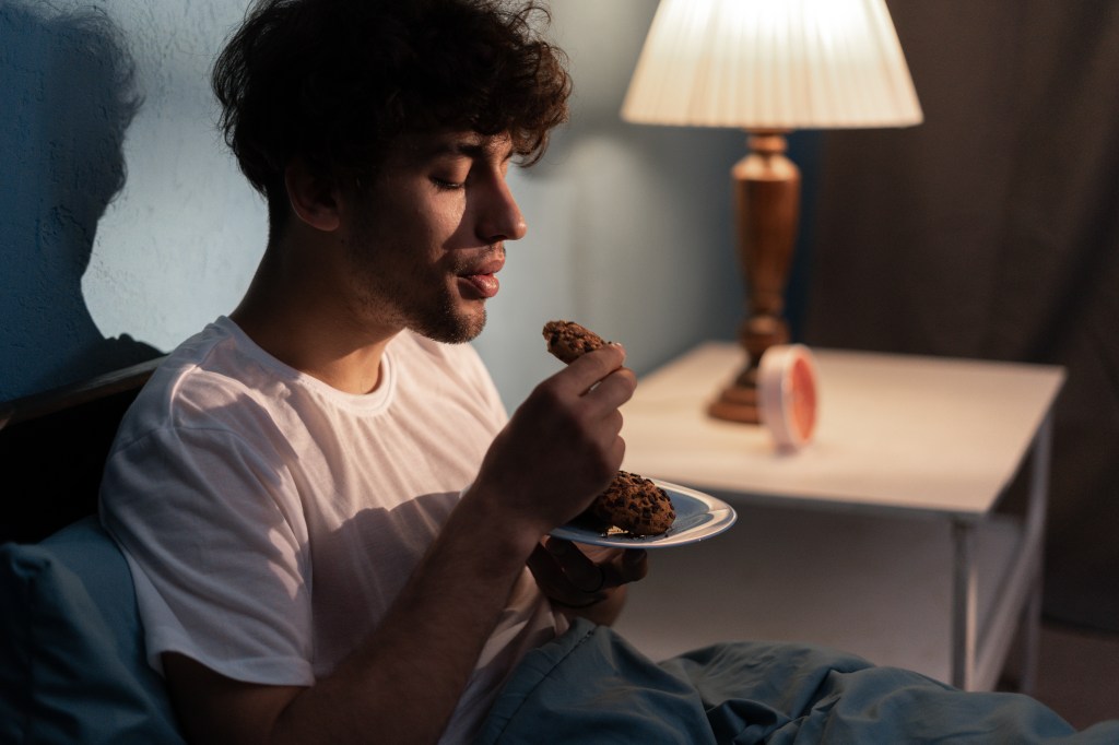 A tired young man eats cookies in bed at night.