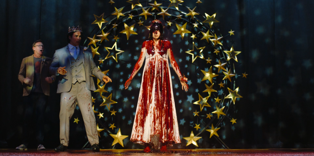 A young woman stands on stage drenched in blood, flanked by a shocked man in a crown and suit, with a large star-studded arch behind them.