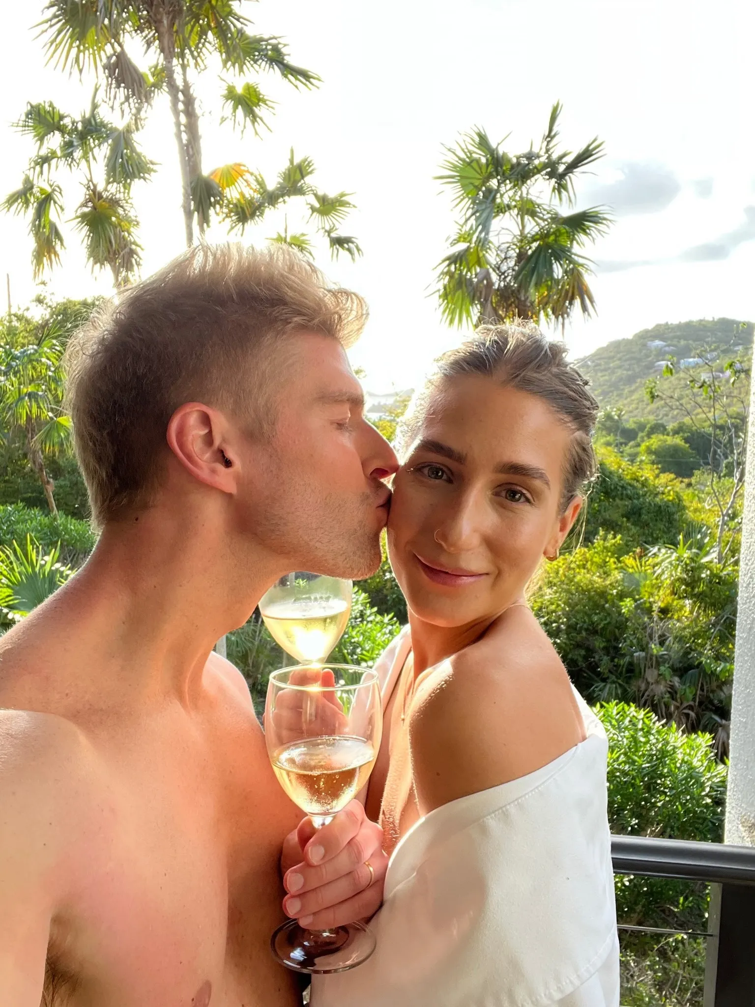 Kyle Cooke kisses Amanda Batula on the cheek as they hold up glasses of wine outdoors on their honeymoon.