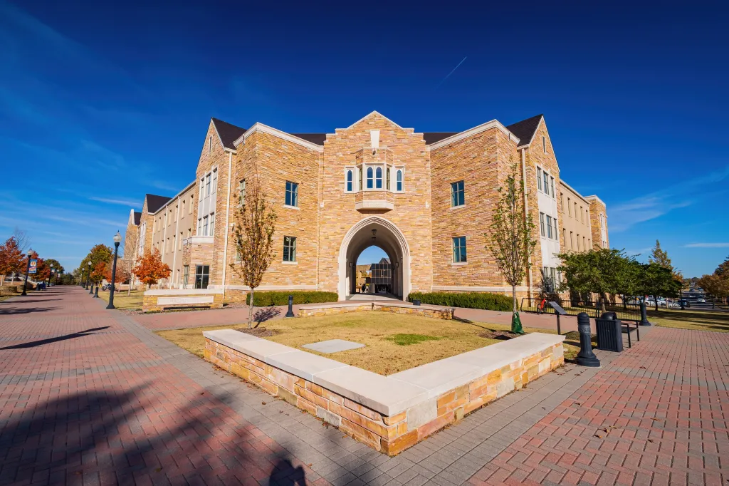 A sunny view of Hardesty Hall on the campus of The University of Tulsa, featuring a large stone building with a central arched entryway under a clear blue sky.