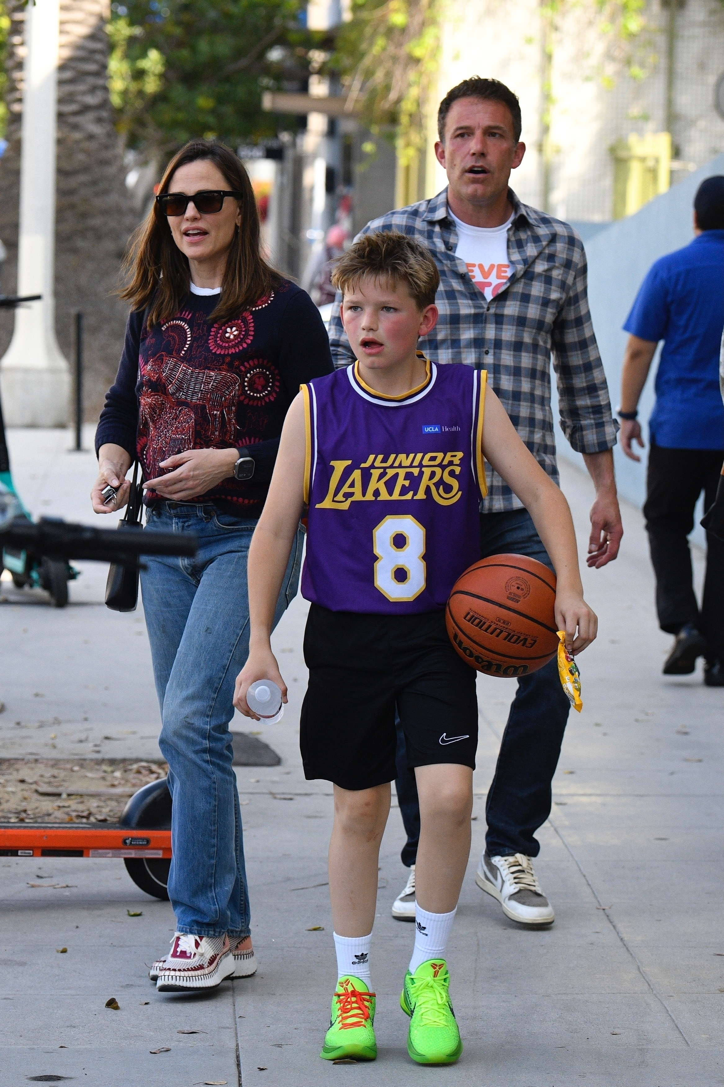 Ben Affleck and Jennifer Garner walk behind their son Samuel, who holds a basketball and a water bottle in a Junior Lakers uniform.
