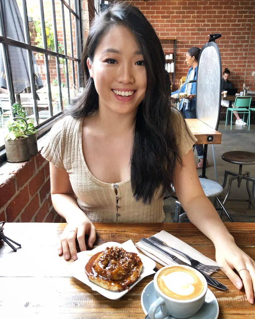 A smiling young woman sits at a wooden table inside Sycamore Kitchen, with a pastry and a latte in front of her.