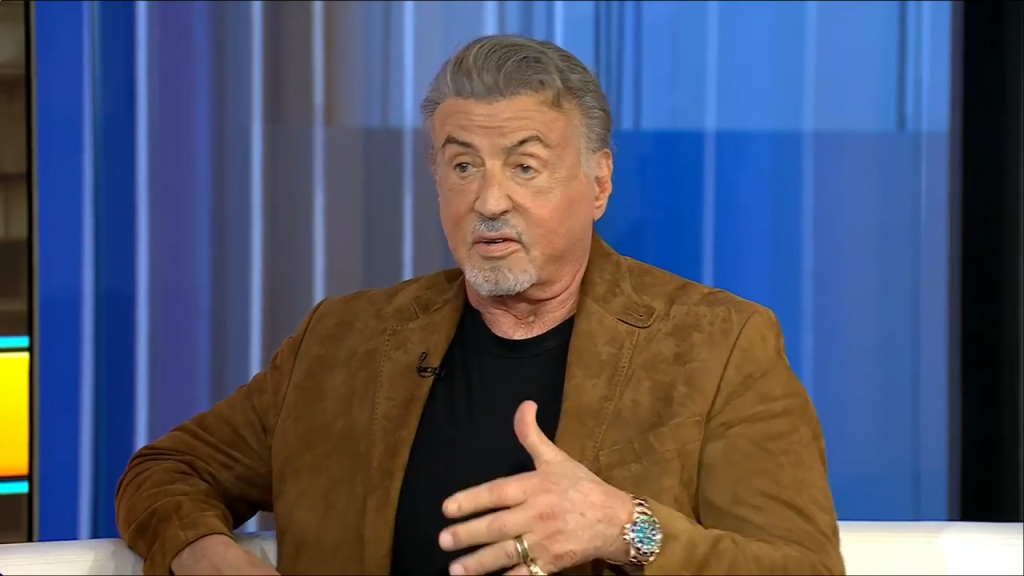 Sylvester Stallone sitting in a brown jacket on a set with a blue background.