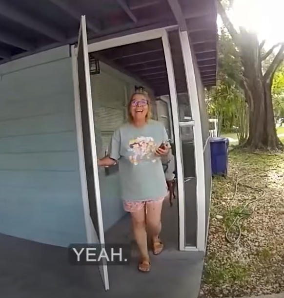 A smiling woman walks through an open door on a porch, holding a phone.