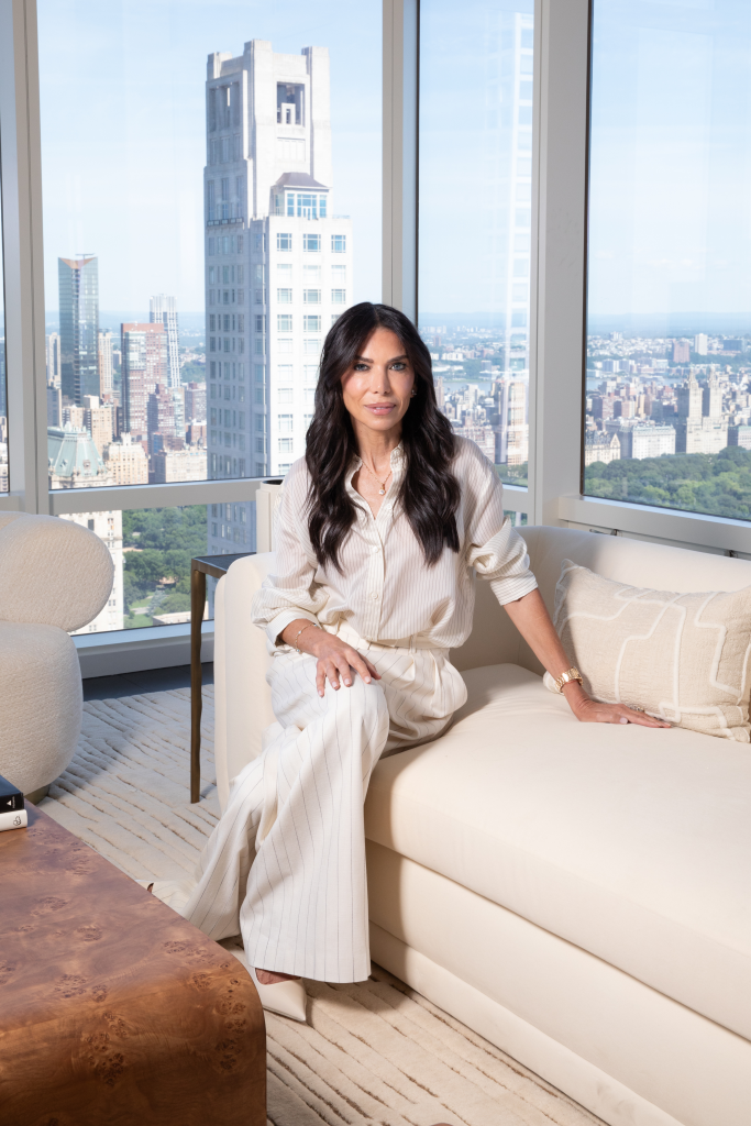 Tanya Zuckerbrot sits on a white couch in her Midtown Manhattan apartment, with floor-to-ceiling windows showing the city skyline behind her.