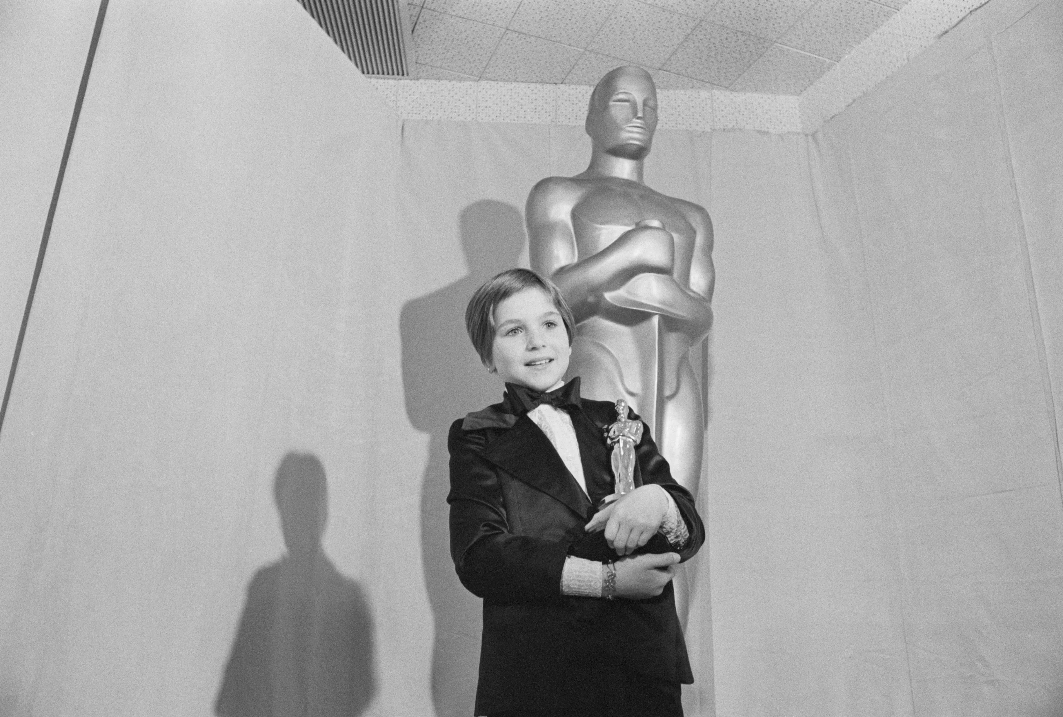 Tatum O'Neal smiling and holding her Oscar statuette in front of a giant Oscar statue.