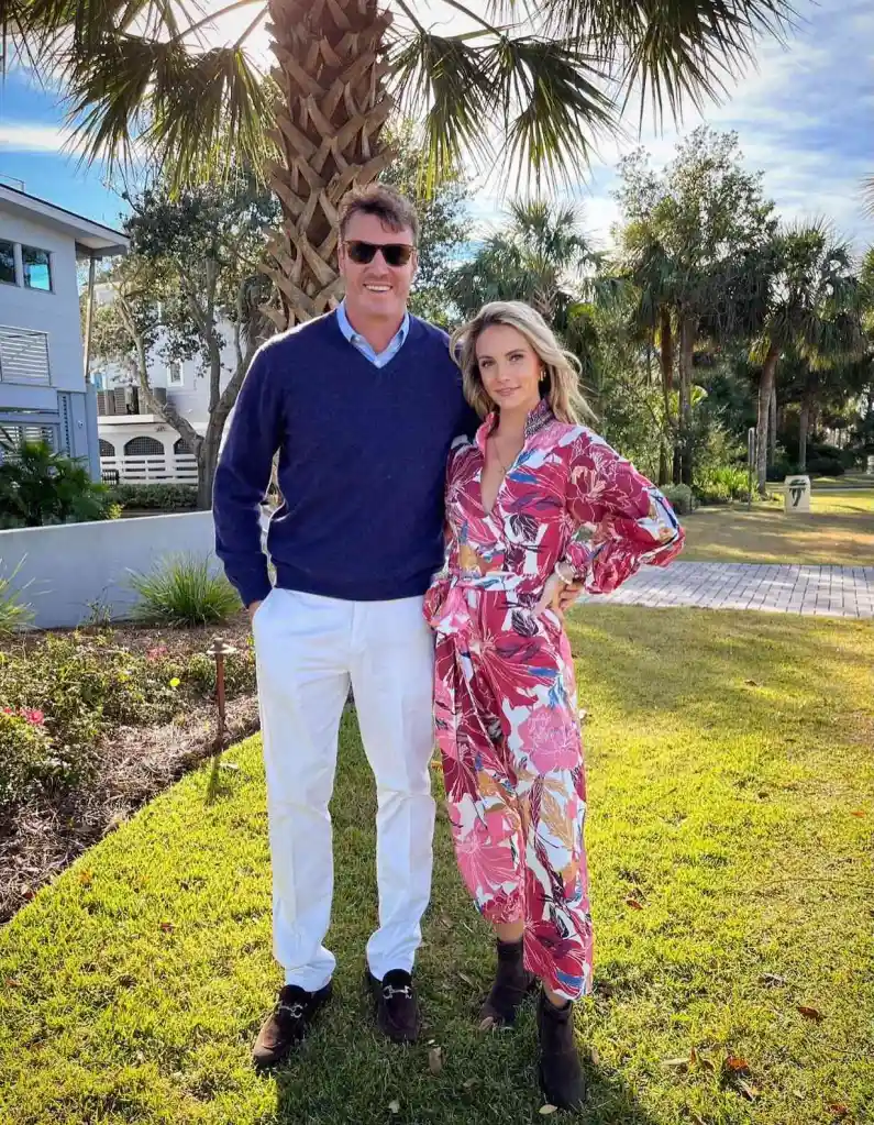 Taylor Ann Green and Shep Rose stand together outdoors, with Taylor wearing a vibrant pink floral dress and Shep in a navy sweater and white pants.