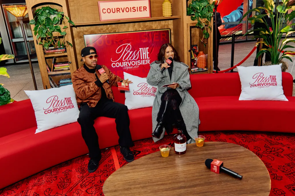 Terrence J and Rocsi Diaz seated on a red couch in an open-air living room, holding microphones and speaking.