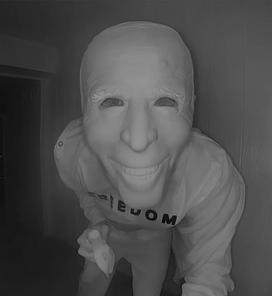 A person wearing a Joe Biden mask leans close to a doorbell camera while holding a banana.