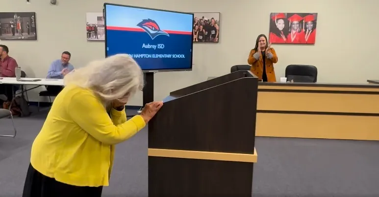 A woman in a yellow cardigan leaning over a podium, overcome with emotion, while people in the background applaud.