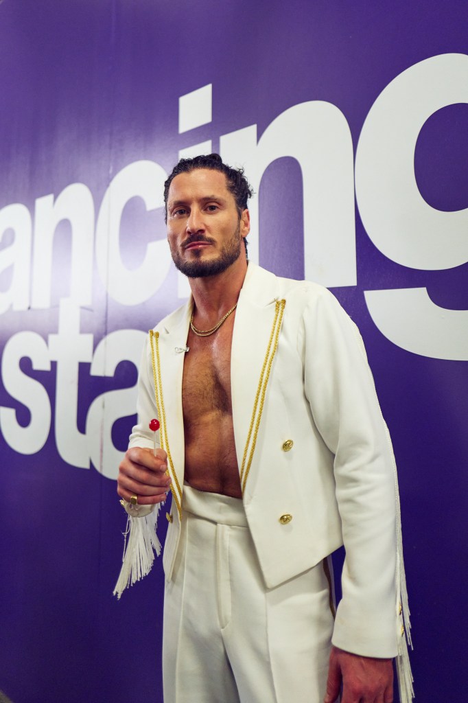 Valentin Chmerkovskiy in a white jacket, holding a lollipop in front of a purple backdrop.