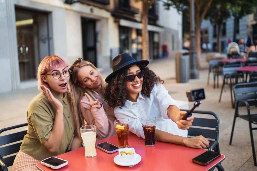 Three women friends at a table taking a photo with a phone. They're young and attractive.