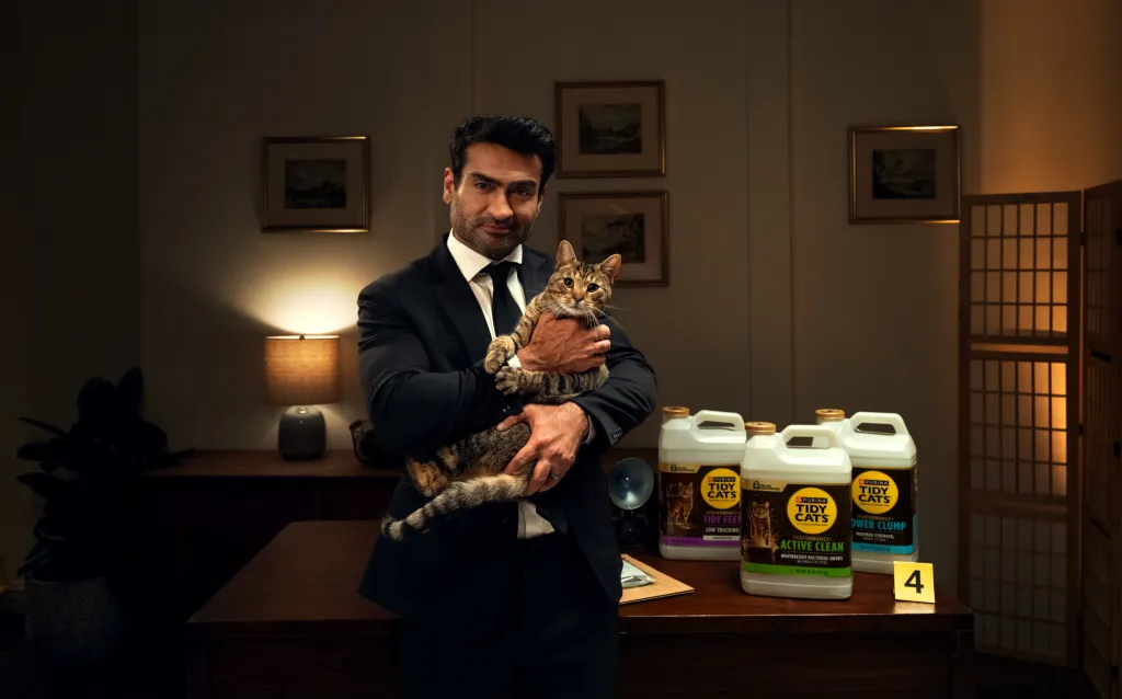 Kumail Nanjiani in a suit holding a tabby cat in an office with Tidy Cats litter containers on the desk.