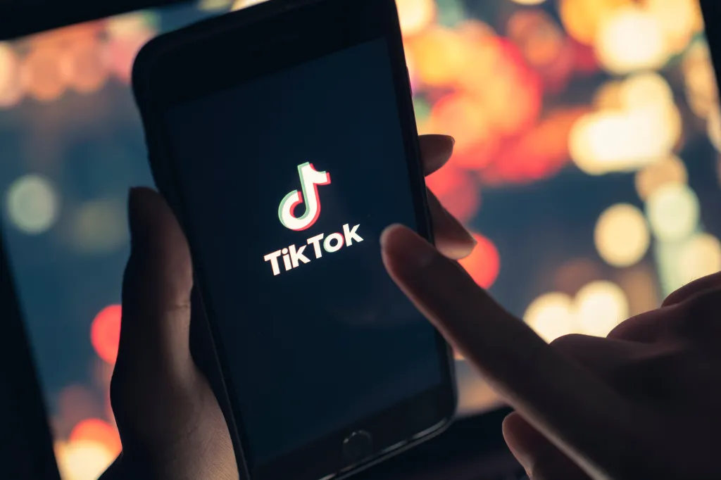 A hand holding a smartphone displaying the TikTok app logo on screen against a blurry bokeh background.