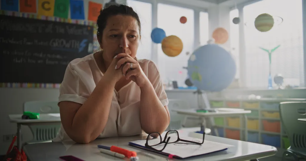 A tired female teacher sitting alone at her desk in an empty classroom, experiencing stress and burnout.
